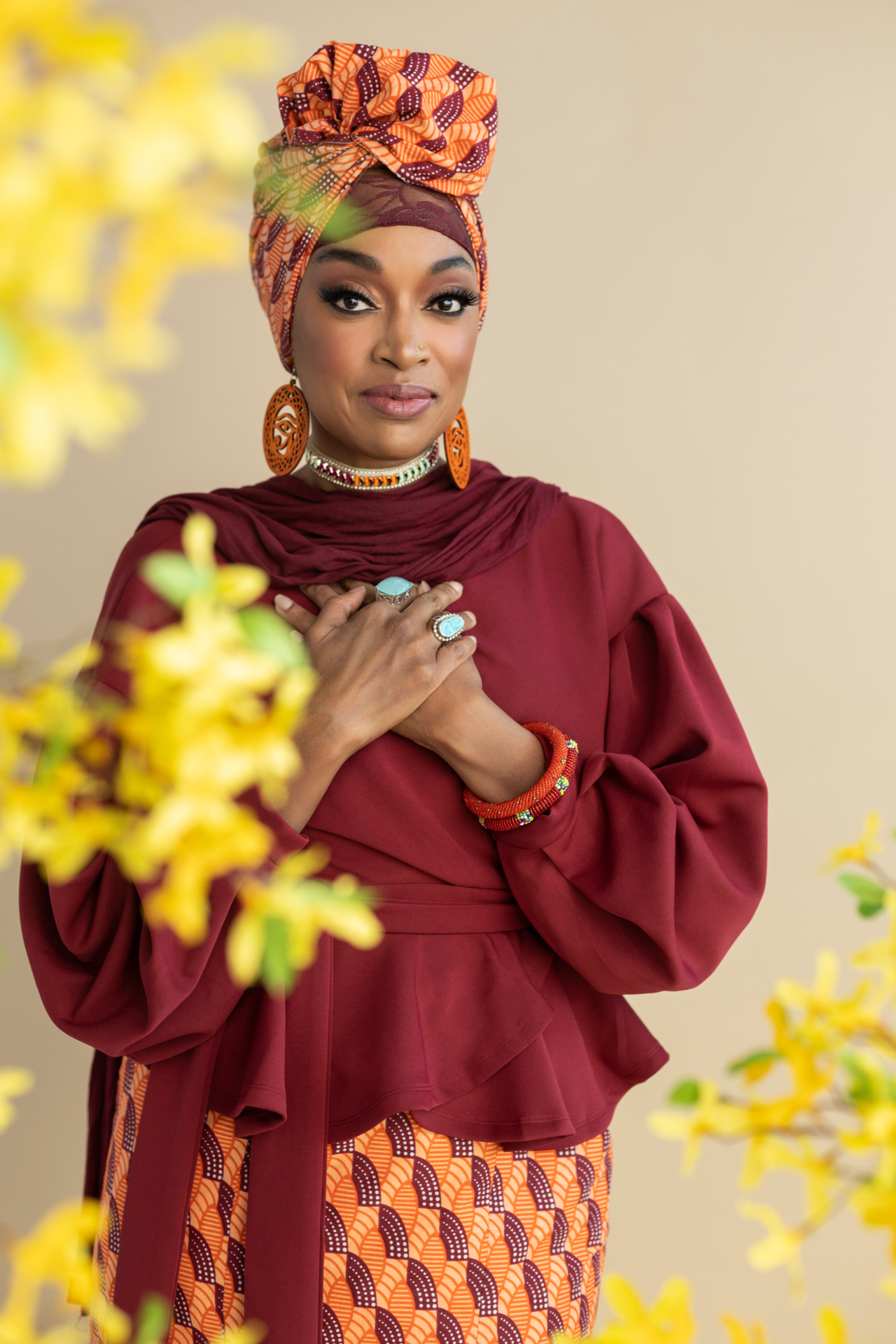 A woman in a burgundy dress and orange headwrap, hands crossed at her heart