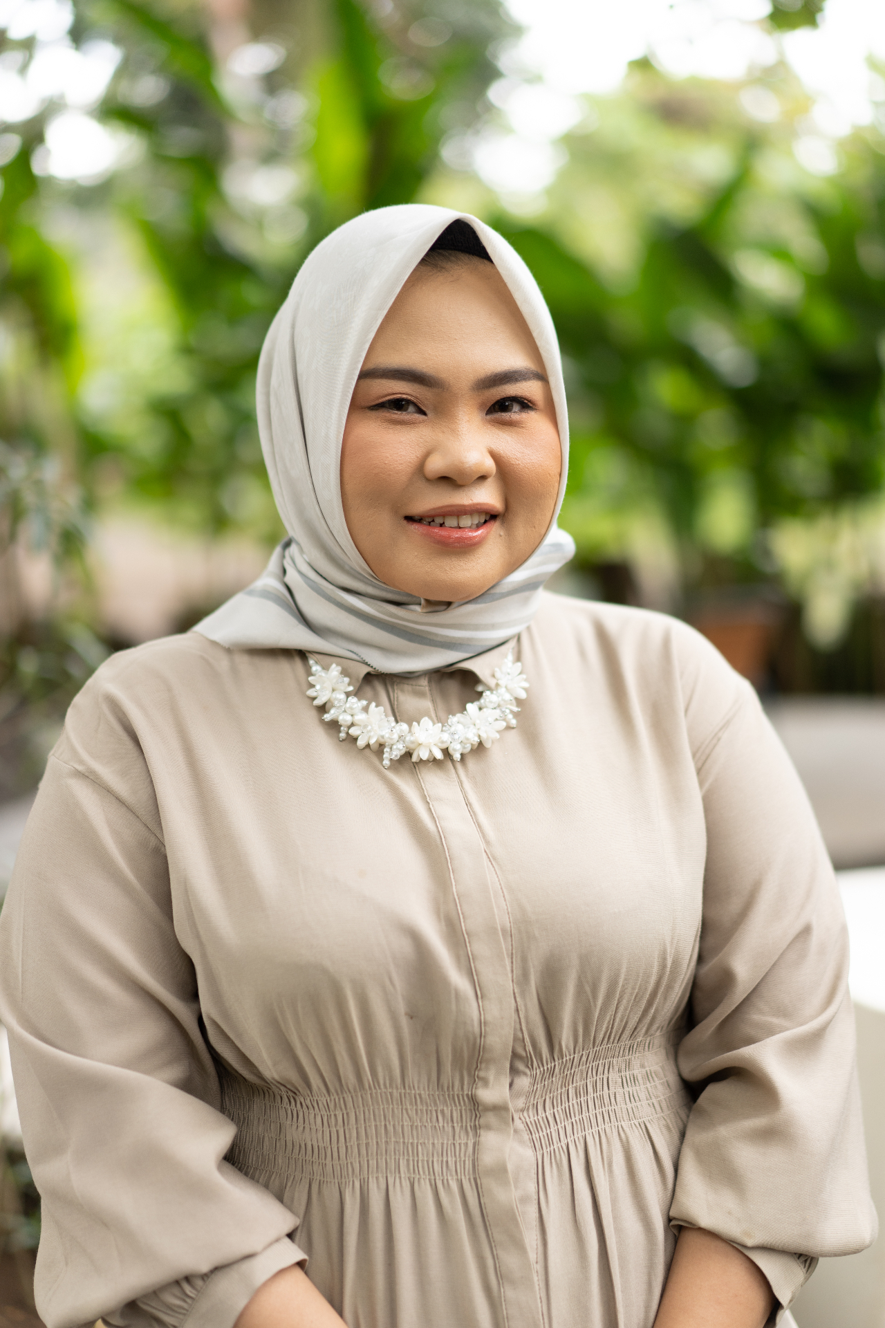 A woman in a soft grey hijab smiling warmly amid green foliage
