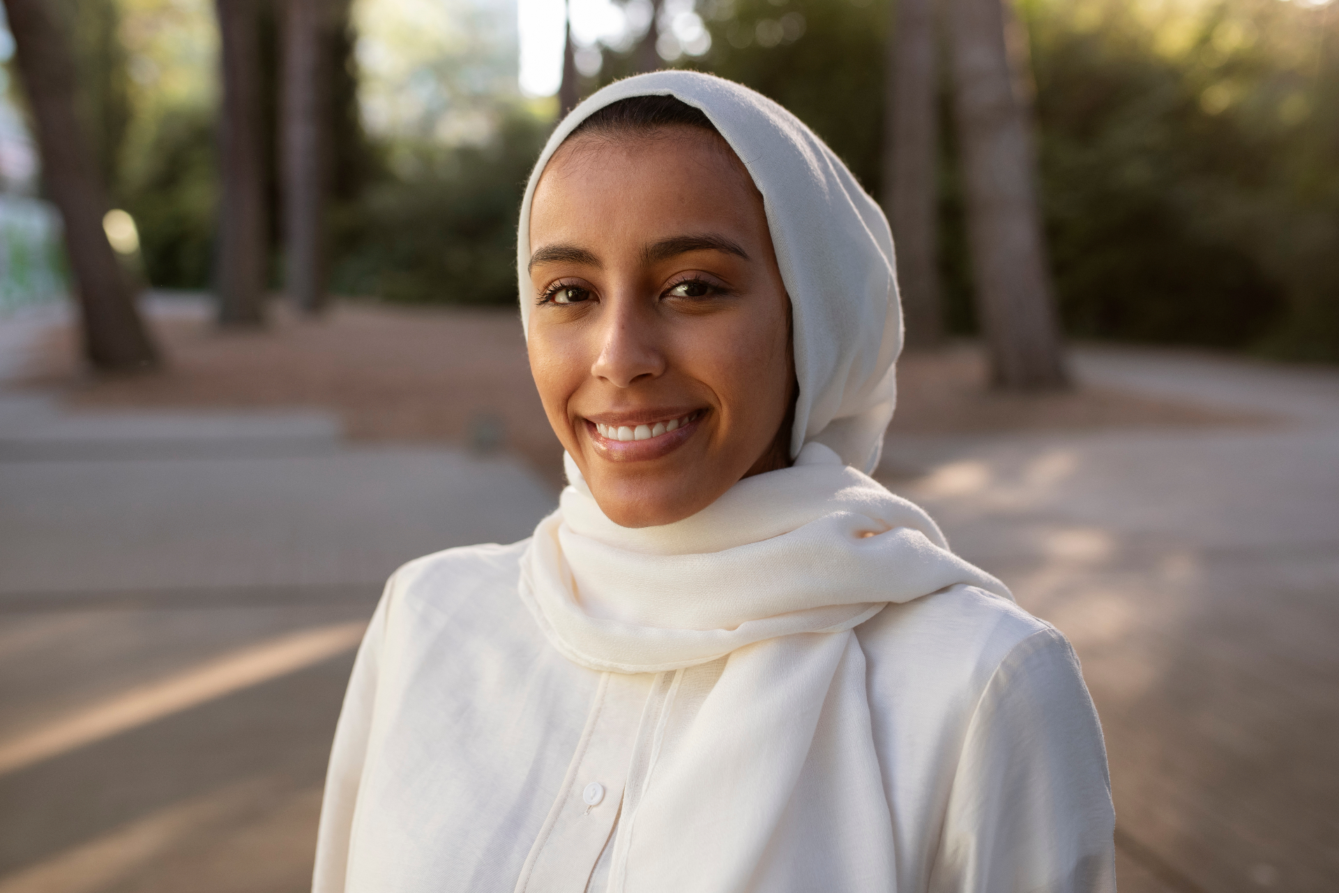 A Muslim woman in a white hijab, smiling warmly outdoors among trees